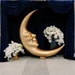 A moon shaped statue with a face next to two stunning flower arrangements.