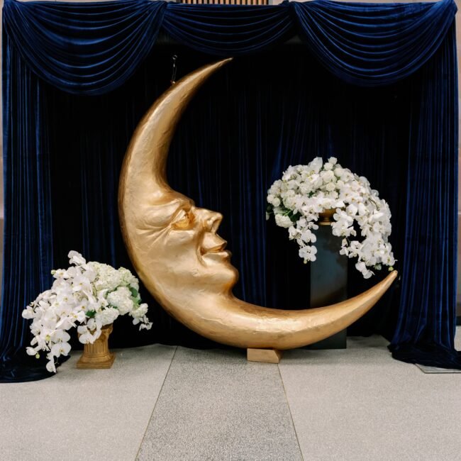 A moon shaped statue with a face next to two stunning flower arrangements.