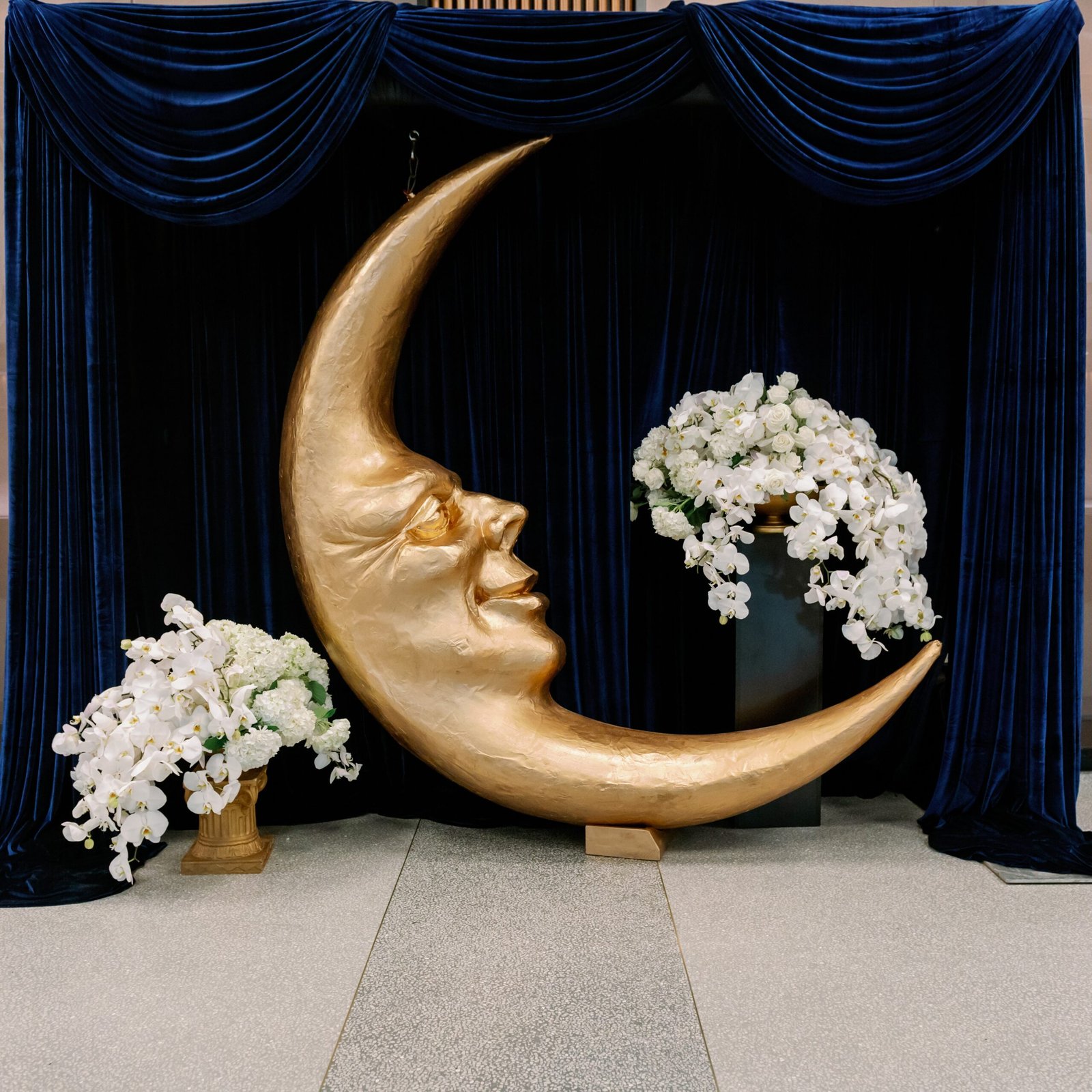A moon shaped statue with a face next to two stunning flower arrangements.