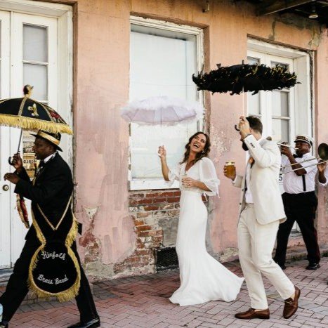 Just-married couple joyfully second lining through the streets of New Orleans, leading the parade with umbrellas and dancing to lively brass band music.