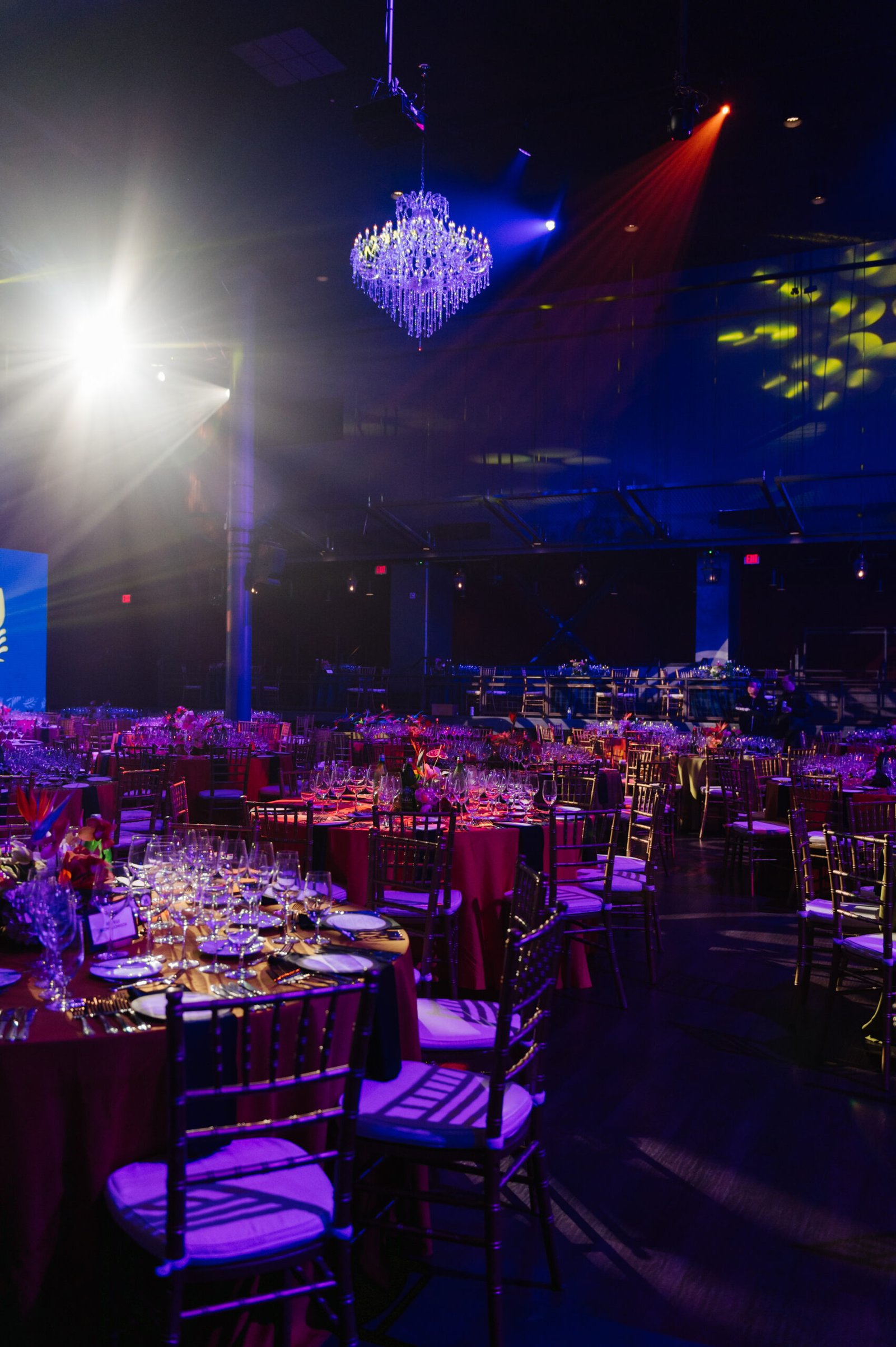 Floral centerpieces and formally set tables beneath a crystal chandelier and colorful event lighting at Carnivale du Vin.