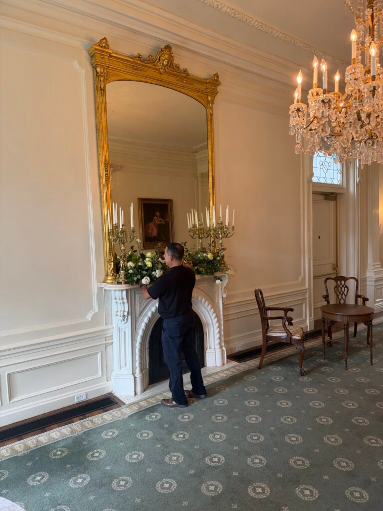 A floral designer arranging white flowers and greenery on a marble mantel beneath an ornate gold mirror.